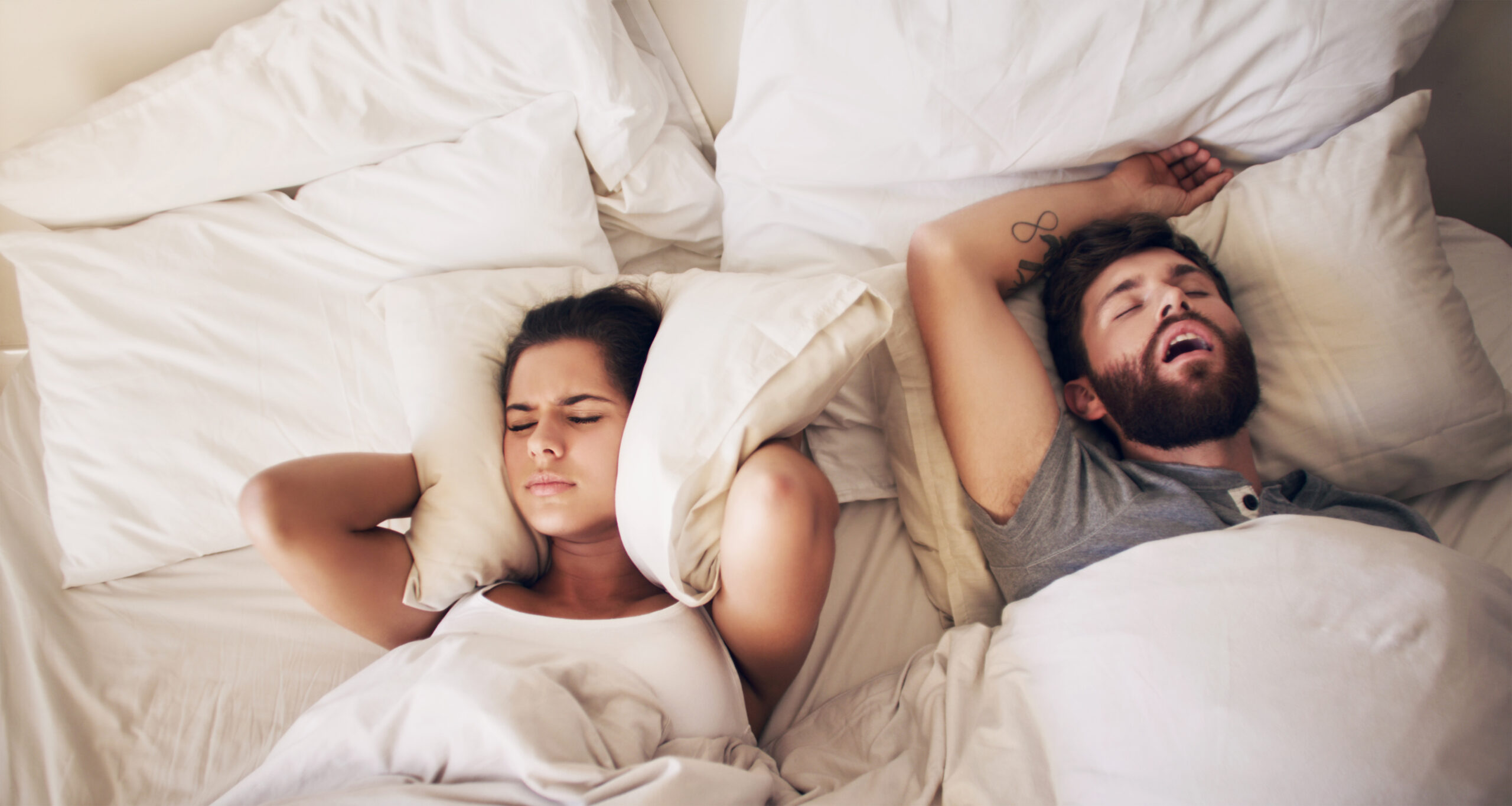 Man snoring in bed while woman covers her ears with pillow, illustrating sleep apnea symptoms treated at Randall K. McVey, DMD PA in Garden City, KS